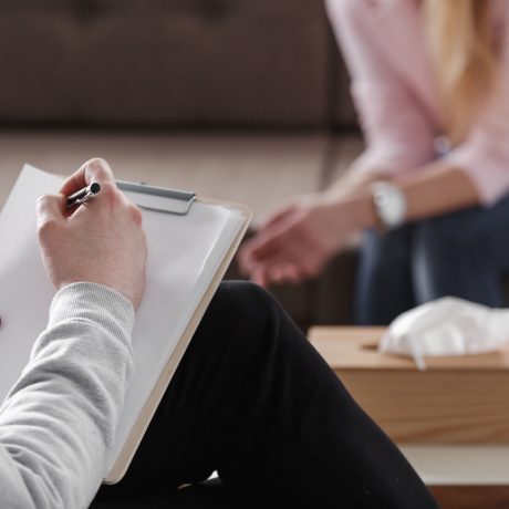 Close-up of therapist hand writing notes during a counseling session with a single woman sitting on a couch in the blurred background.